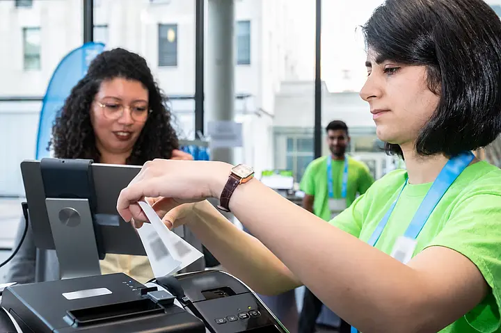 Eine Mitarbeiterin nimmt ein gedrucktes Namensschild aus dem Drucker; persönliche Angaben sind unkenntlich