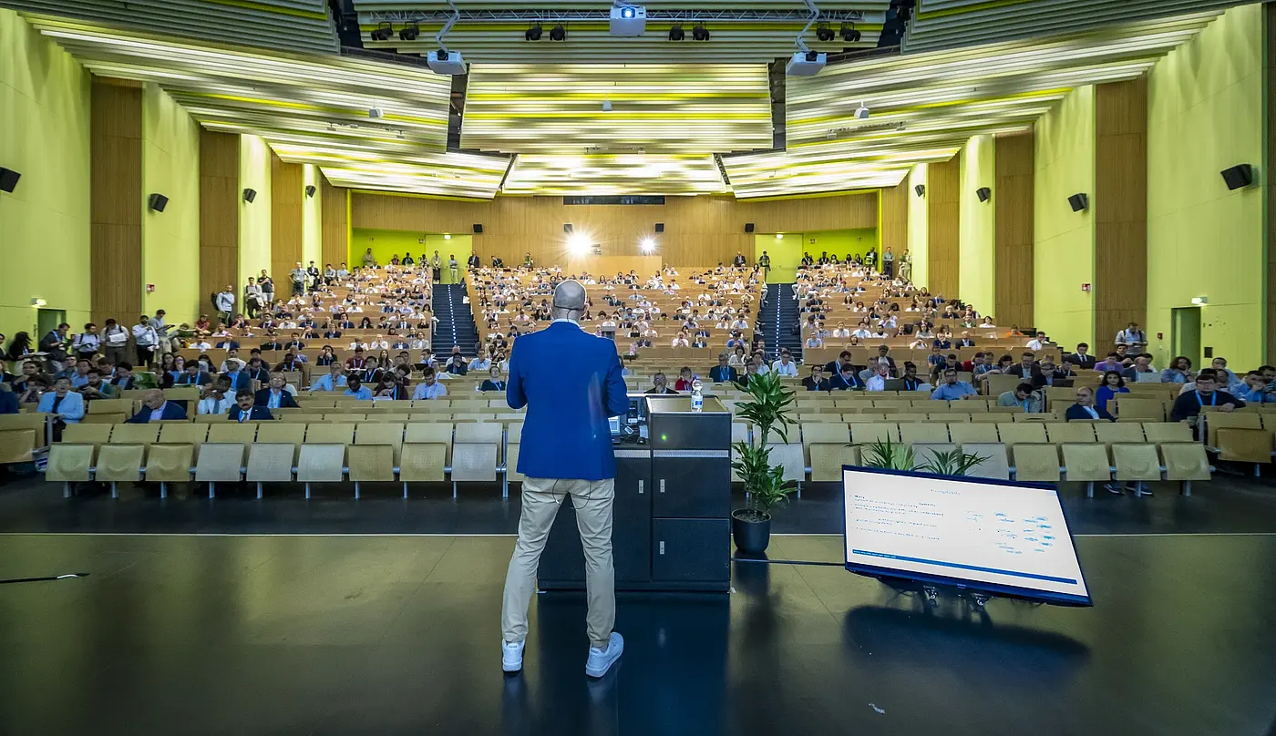 Blick von der Bühne über einen Vortragenden zum Publikum mit Vorschaumonitor am Bühnenrand