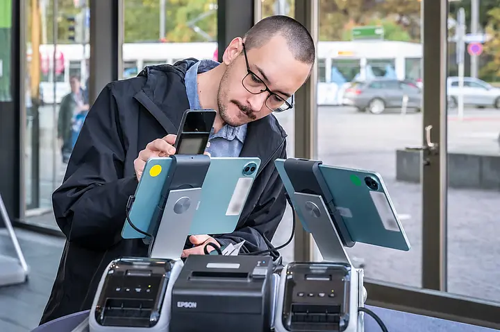 Ein Teilnehmer hält sein Smartphone vor die Kamera eines Check-in-Tablets; persönliche Daten und Codes sind unkenntlich