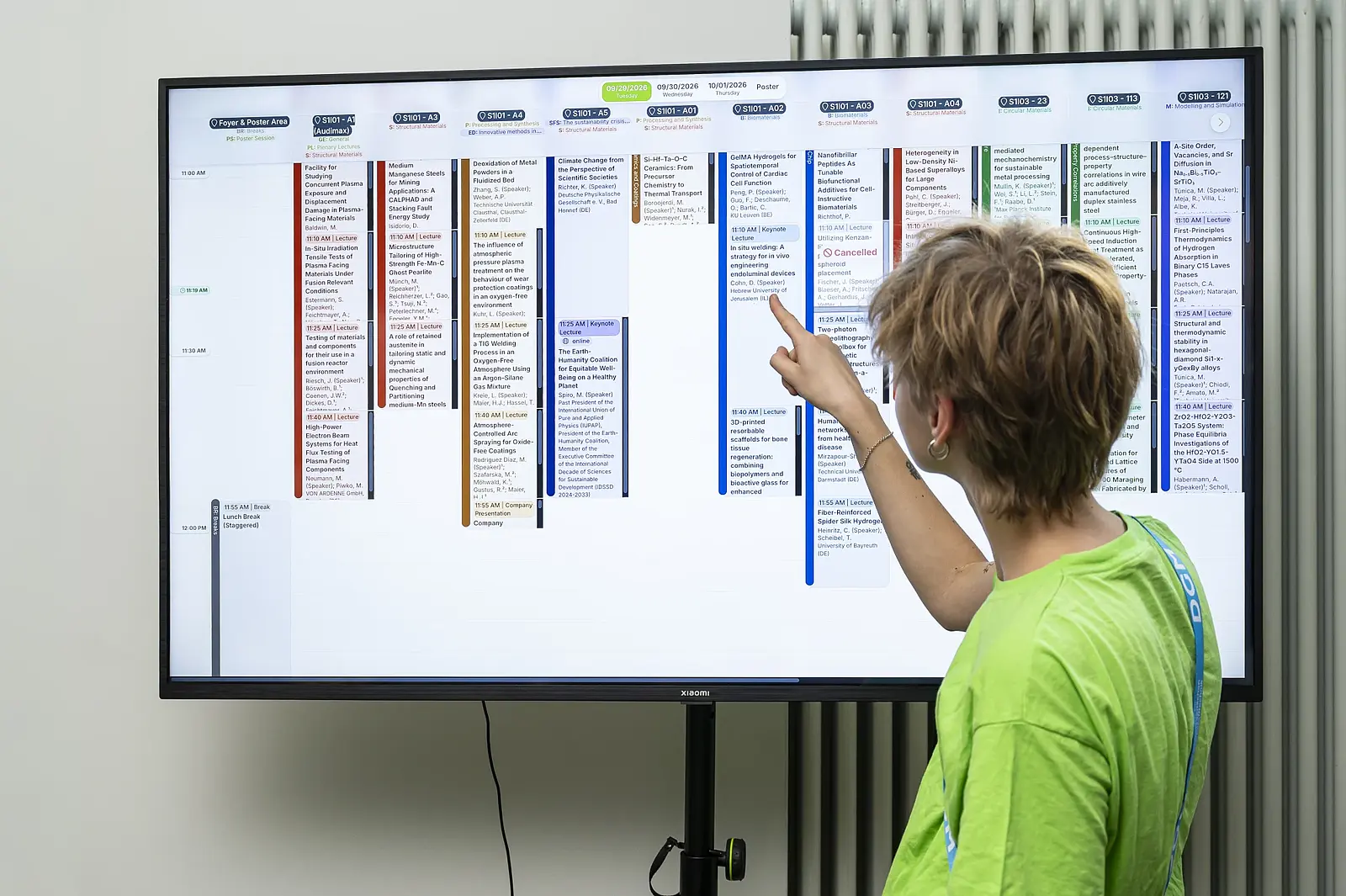 Eine Mitarbeiterin zeigt auf einen Vortrag im interaktiven Konferenzprogramm auf einem großen Display