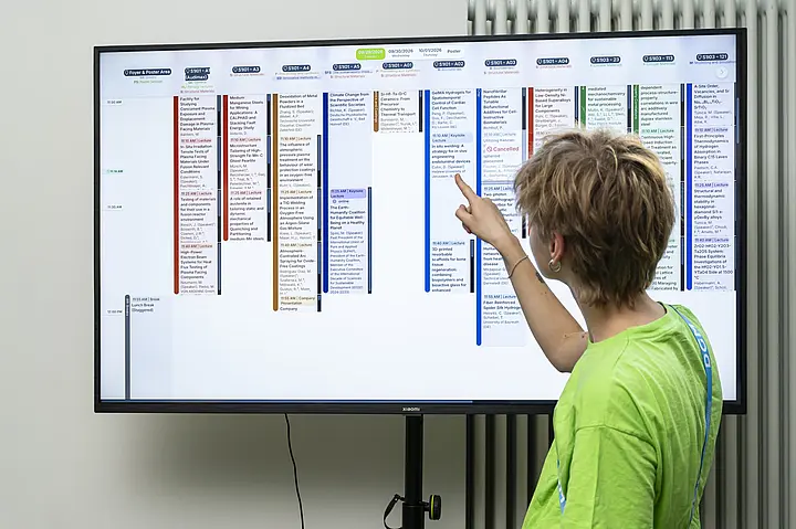Eine Mitarbeiterin zeigt auf einen Vortrag im interaktiven Konferenzprogramm auf einem großen Display